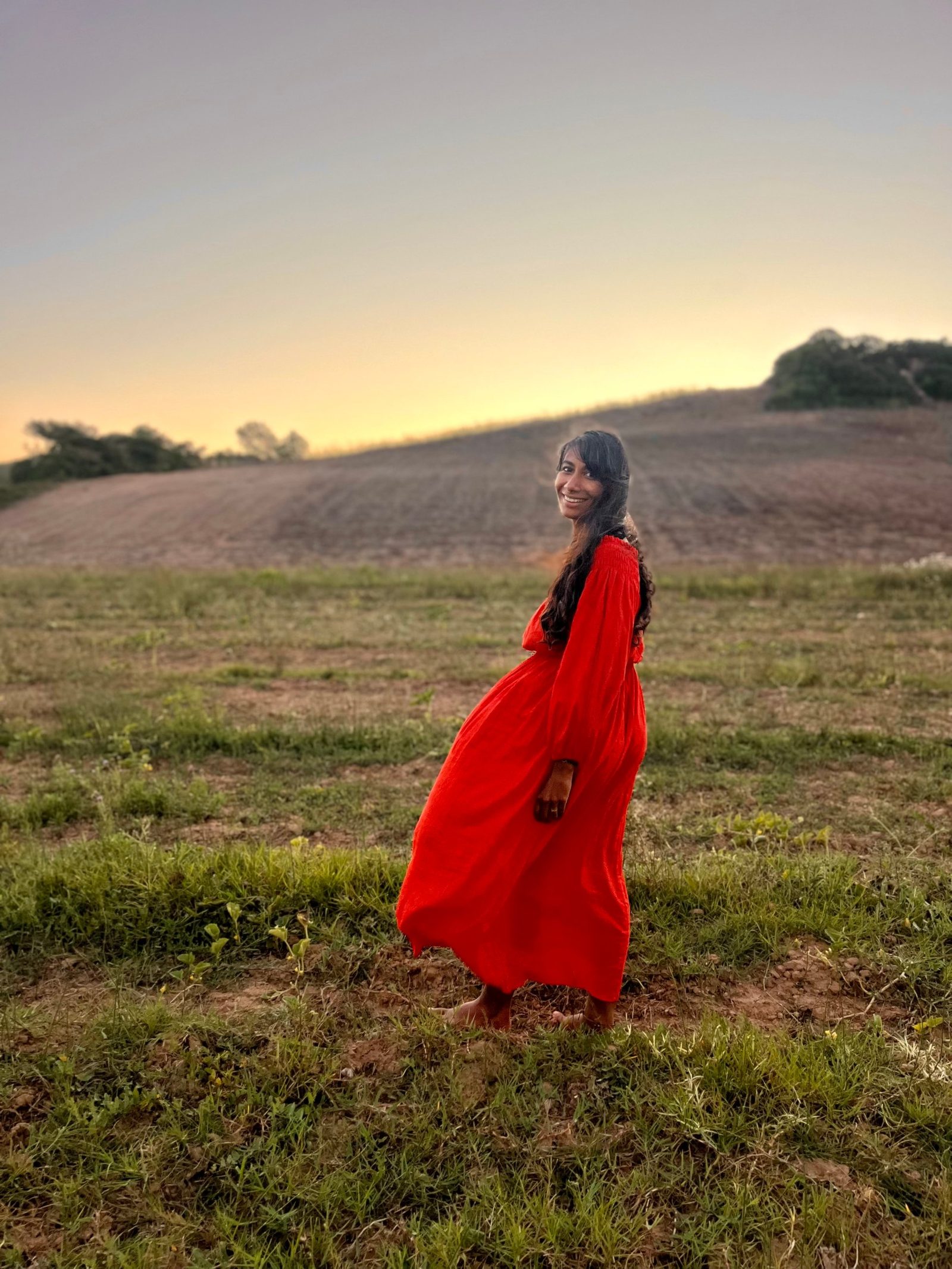 Davina en robe rouge, tournée vers l’objectif, dans un champ à la lumière du coucher de soleil.