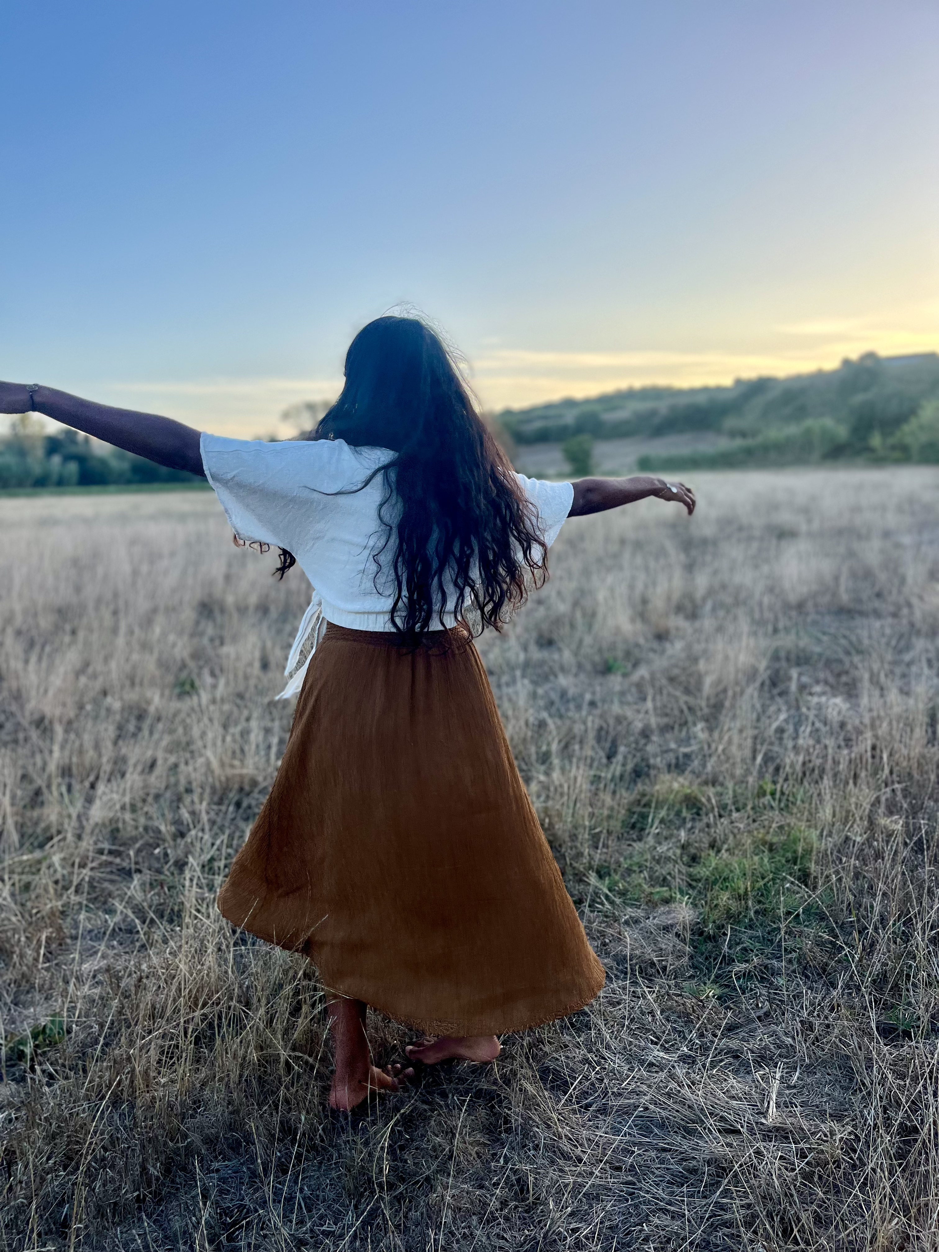 Davina Charlène de dos, les bras ouverts dans un champ au coucher du soleil