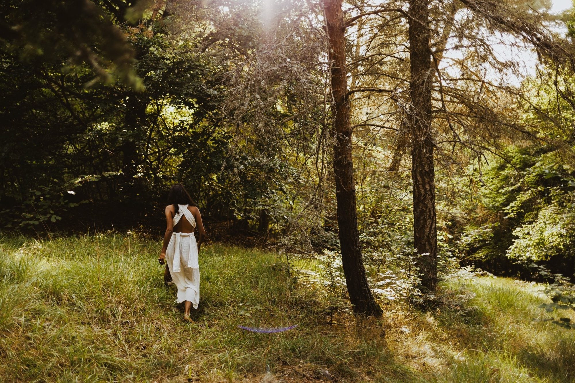 Davina de dos, en robe blanche, qui marche dans l’herbe vers la forêt