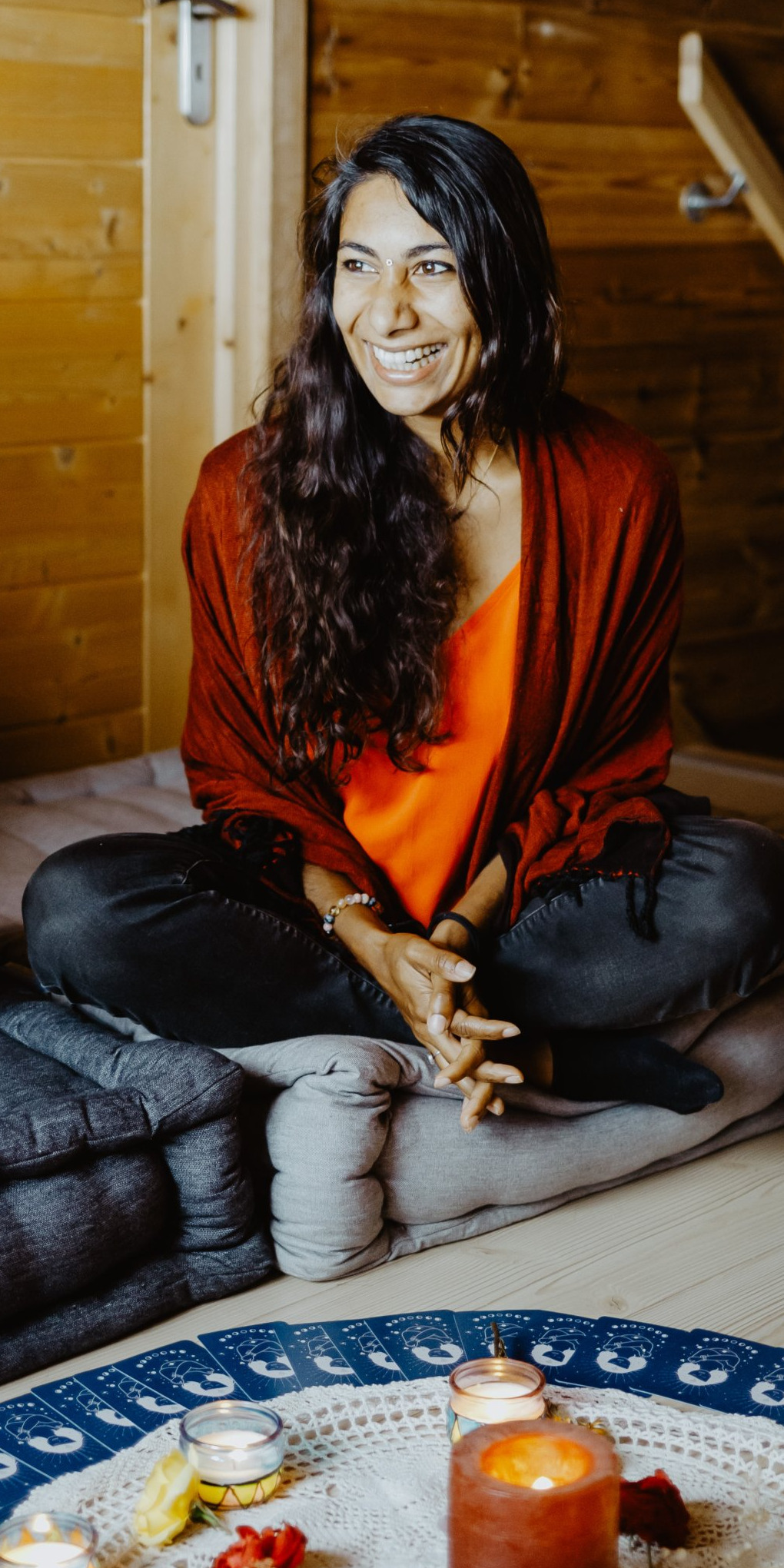 Davina, assise en tailleur et souriante, un châle rouge sur les épaules, devant un cercle de cartes et des bougies allumées.