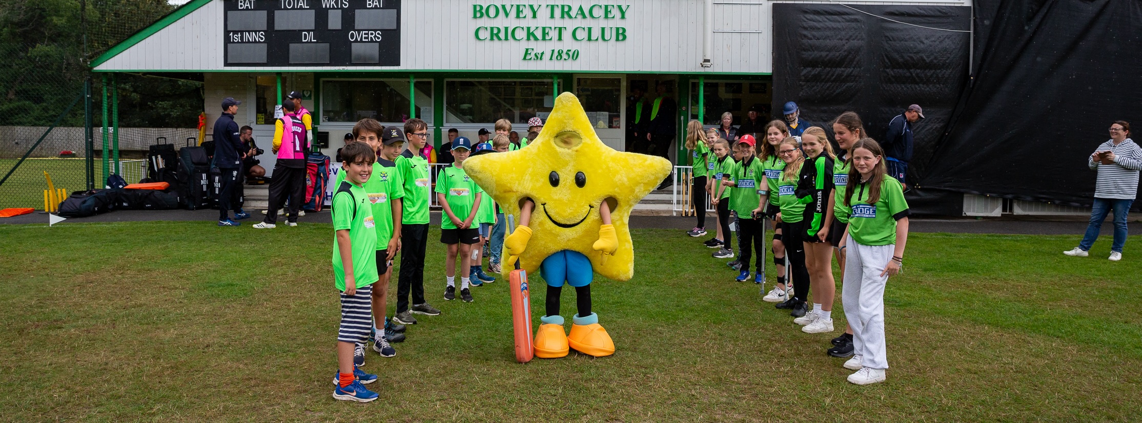 Boys and girls cricketers in front of a cricket pavilion with a all star mascot