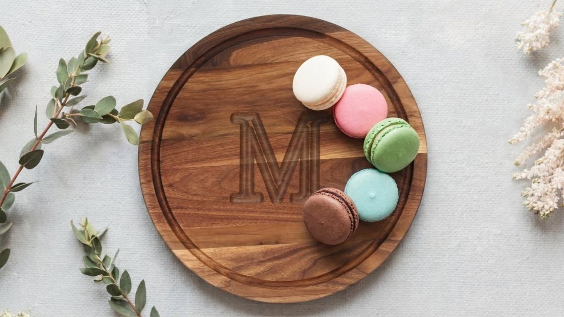 op-down view of a round Monogrammed Walnut Cutting Board featuring the letter 'M', styled with colorful macarons and eucalyptus on a white surface.