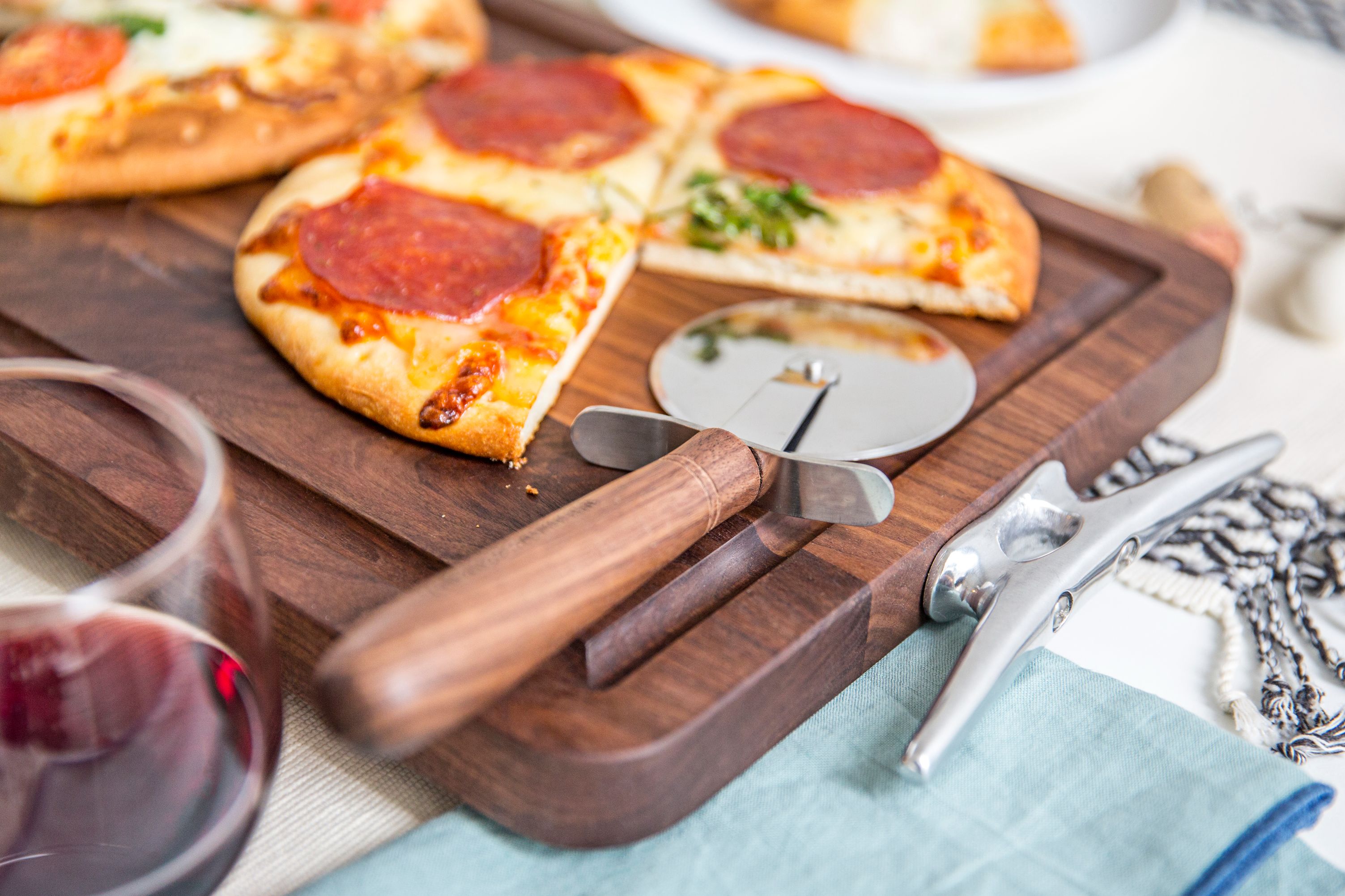 A detailed close-up of a sliced pepperoni flatbread pizza on a custom dark walnut cutting board, highlighting its deep juice groove and rich wood grain. A matching walnut-handled pizza cutter rests on the board next to a glass of red wine and a modern corkscrew.