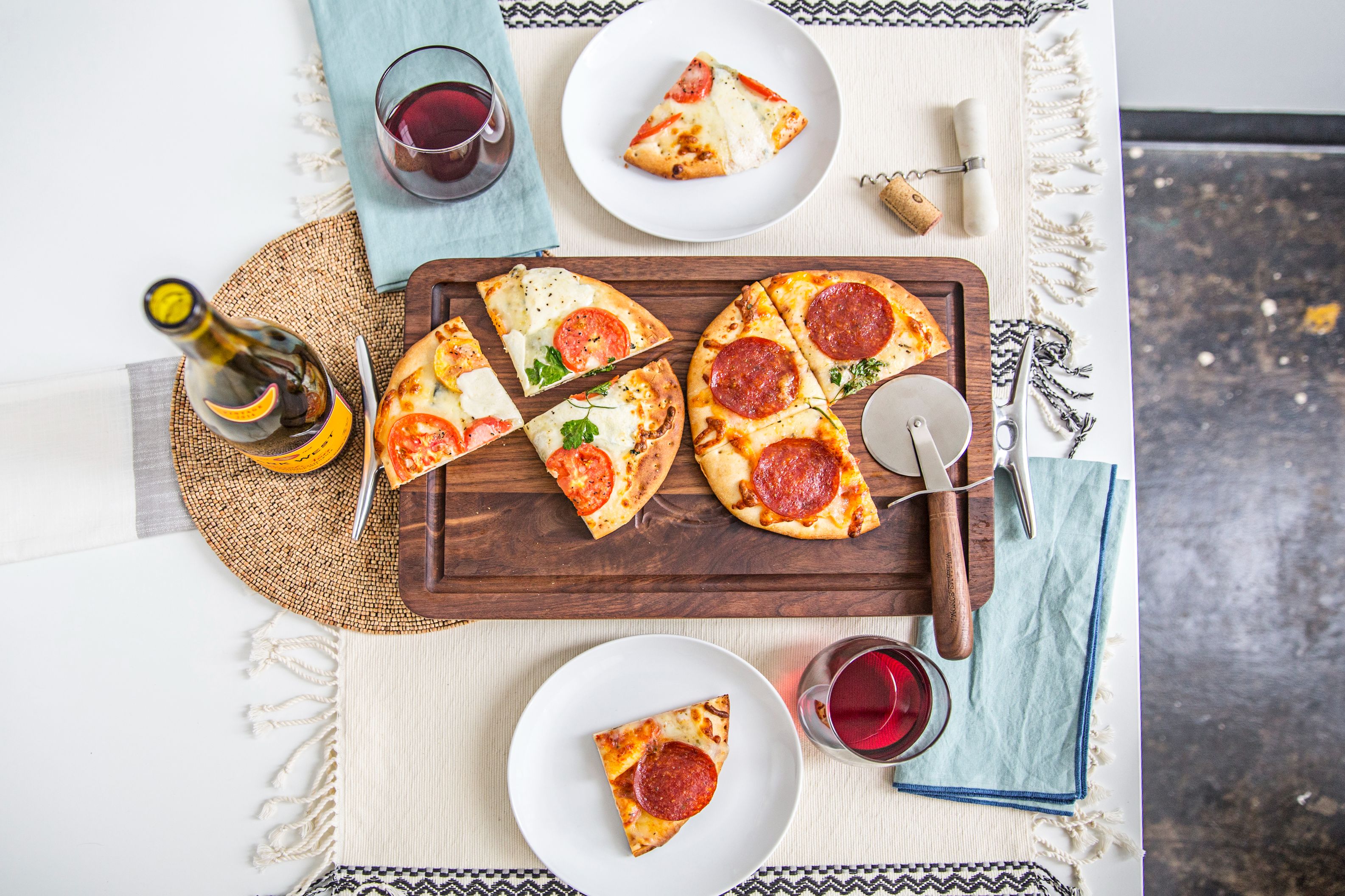 n elegant pizza night scene featuring two artisanal flatbread pizzas on a custom dark walnut cutting board with a juice groove, styled with wine, glasses, and a pizza cutter on a clean white table setting.