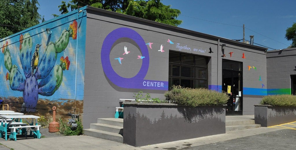 Photo of the PDX Q Center building. The building is mosttly gray with a colorful mural on the left wall featuring a cactus and bird. On the front wall is another mural featuring a large purple 