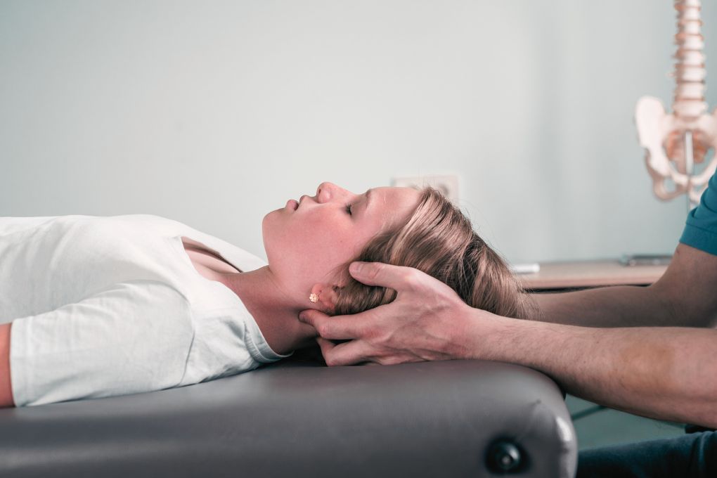 Eine Frau liegt entspannt auf einer Behandlungsliege, während ein Therapeut ihre Nackenmuskulatur mit den Händen sanft massiert – eine mögliche Unterstützung bei Stress, Anxiety und Verspannungen in den Wechseljahren. Im Hintergrund ist ein Modell der Wirbelsäule zu sehen.