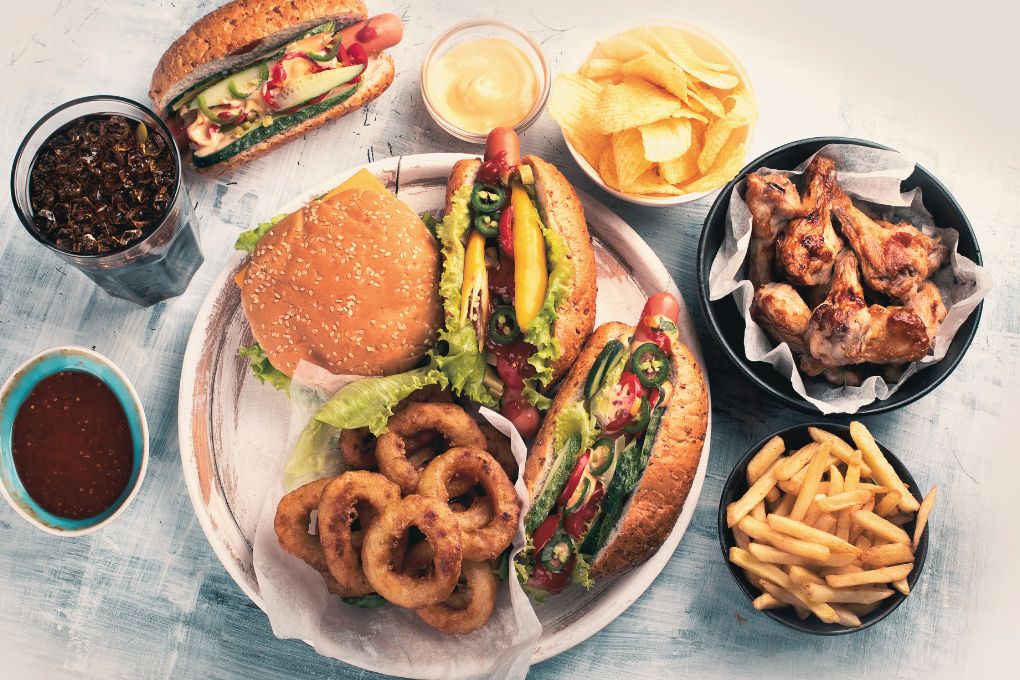 Eine Auswahl an ultrahochverarbeiteten Lebensmitteln, darunter Burger, Hot Dogs, Pommes, Chips, Zwiebelringe, Chicken Wings und Softdrink. Symbolbild für Fast Food und hochverarbeitete Lebensmittel, die mit ungesunder Ernährung und Gewichtszunahme in Verbindung stehen.