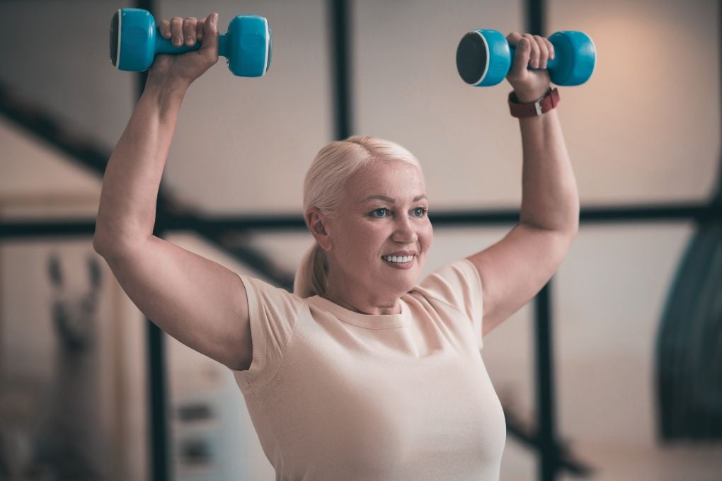Eine ältere Frau mit heller Haut und weißen Haaren hebt lächelnd Hanteln über den Kopf. Dieses Bild zeigt die Bedeutung von Krafttraining in der Menopause, um Muskelschwund (Sarkopenie) und Knochendichteverlust zu verhindern.