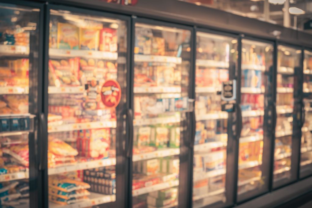 Verschwommene Aufnahme eines Supermarkt-Gefrierschranks mit verschiedenen verarbeiteten Lebensmitteln wie Tiefkühlpizza, Fertiggerichten und Snacks. Symbolbild für ultrahochverarbeitete Lebensmittel (Ultra-Processed Foods) und ihre Verbreitung im Einzelhandel.