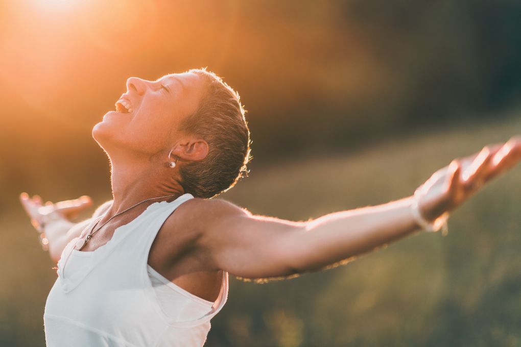Frau mittleren Alters steht mit ausgebreiteten Armen in der Natur und genießt die Sonne. Symbolisiert Freiheit, Lebensfreude und ein positives Lebensgefühl während der Wechseljahre.