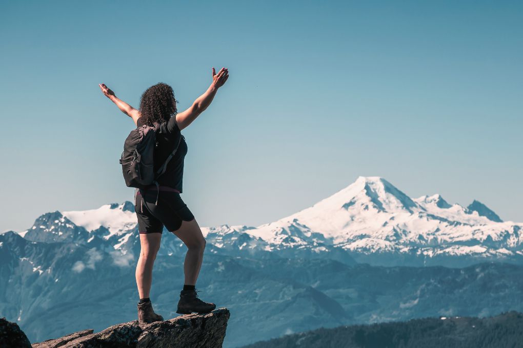 Eine Frau steht mit ausgebreiteten Armen auf einem Berggipfel und blickt auf eine schneebedeckte Berglandschaft – ein Symbol für Stärke, Freiheit und einen aktiven Lebensstil zur Prävention von Osteoporose.