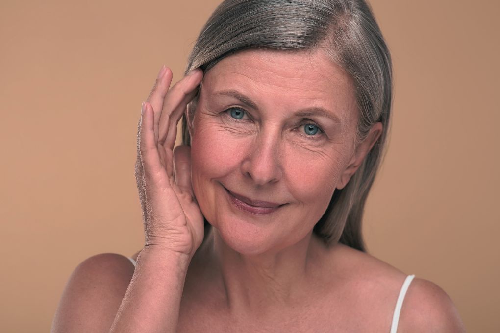 Porträt einer lächelnden älteren Frau mit grauem Haar, die ihre Hand sanft an ihr Gesicht legt. Ihre Haut zeigt natürliche Zeichen des Alterns wie feine Linien und Fältchen. Das Bild unterstreicht das Thema Hautveränderungen in den Wechseljahren und die Bedeutung von Selbstfürsorge und Pflege.