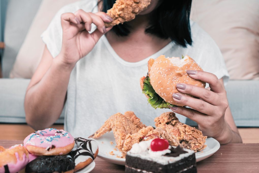 Frau in weißem Shirt isst einen Burger und frittiertes Hühnchen, vor ihr auf dem Tisch liegen Donuts, Kuchen und weitere ultrahochverarbeitete Lebensmittel. Symbolbild für ungesunde Ernährung, Heißhunger und den Einfluss von Fast Food auf die Gesundheit in den Wechseljahren.