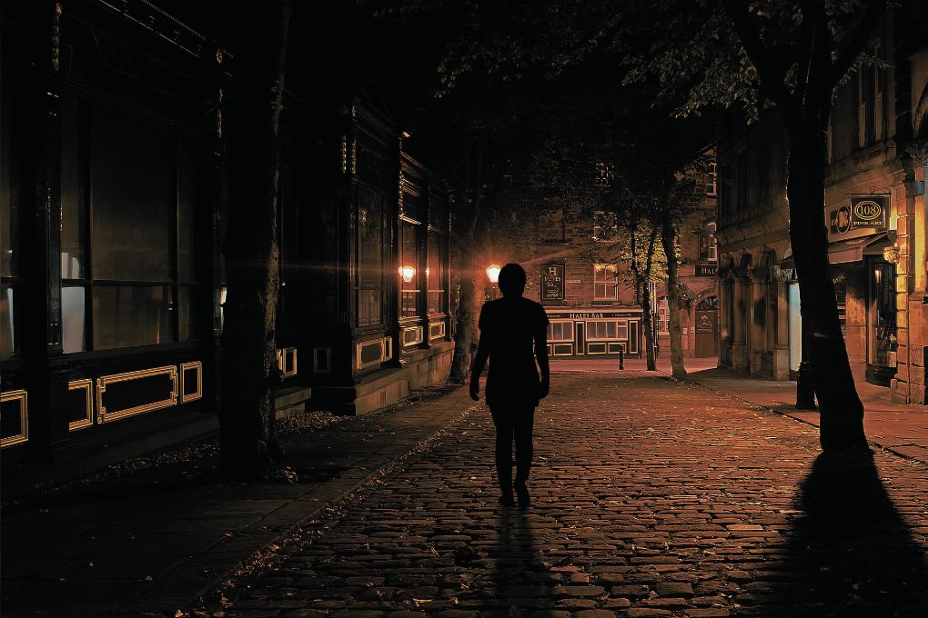 Silhouette einer Person, die nachts langsam auf einem Kopfsteinpflaster in einer schummrig beleuchteten, menschenleeren Straße geht, umgeben von alten Gebäuden und Bäumen – eine Darstellung von Anxiety in den Wechseljahren, verbunden mit Angst, innerer Unruhe und Sehstörungen, wie sie in der Perimenopause auftreten können.
