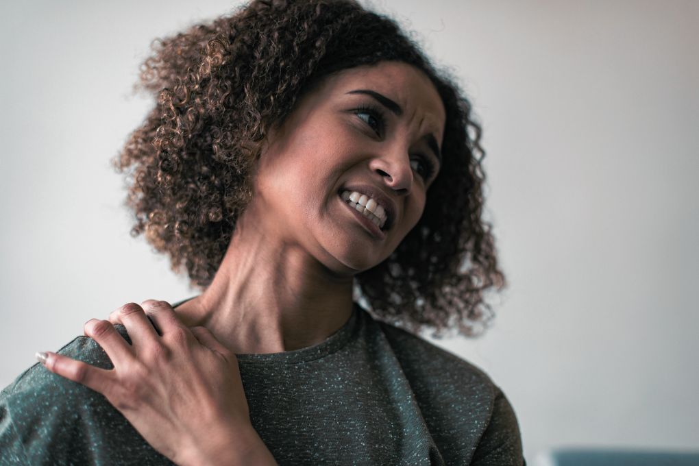Eine Frau hält sich die Schulter und zeigt ein schmerzverzerrtes Gesicht, was auf eine Frozen Shoulder oder muskulär-skelettale Schmerzen hindeuten könnte, die im Zusammenhang mit hormonellen Veränderungen stehen könnten.