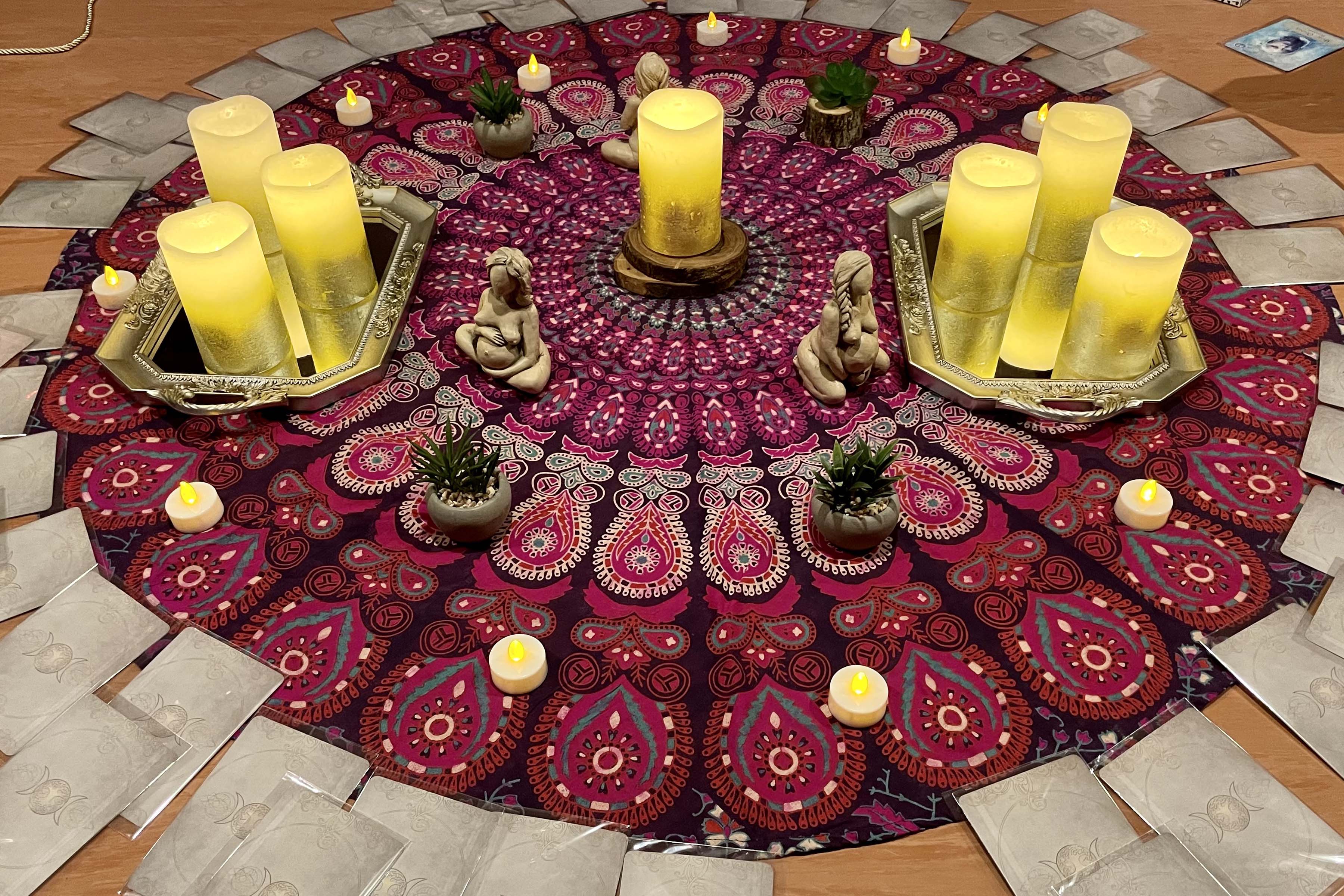 Lana has created a beautiful Sistership Circle shown here with a mandala print tapestry on the floor, surrounded by goddess oracle cards, candles, plants, and statuettes.