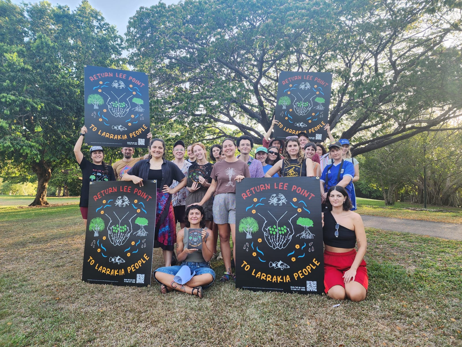 A group of 20 volunteers holding colourful signs calling for Lee Point to be returned to Larrakia people. They are standing on a field in front of a beautiful and lush green tree. The sun is shining and people are smiling. The vibe is warm