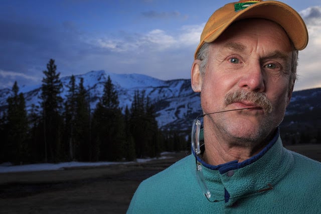 portrait of man in ballcap standing in front of snowy mountains, glasses hanging from corner of mouth