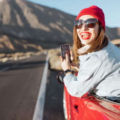 Woman seated in a car on a sunny Tenerife road, smiling at the camera while holding her smartphone displaying navigation directions with Tenerife itinerary.